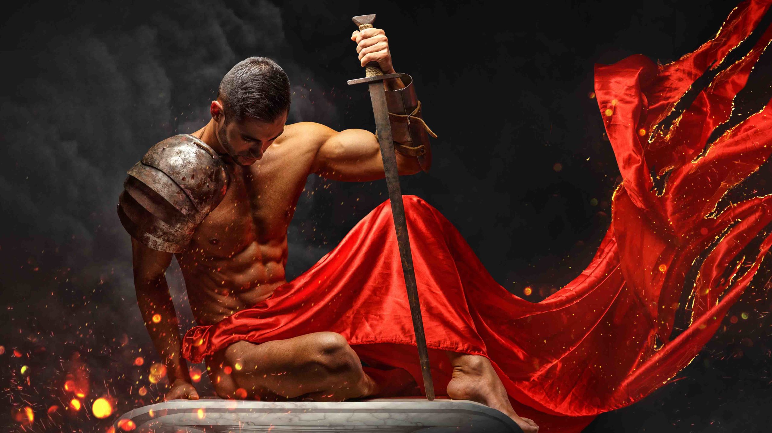 Artistic portrait of muscular male in red waving fabric with fire sparks holding sword.